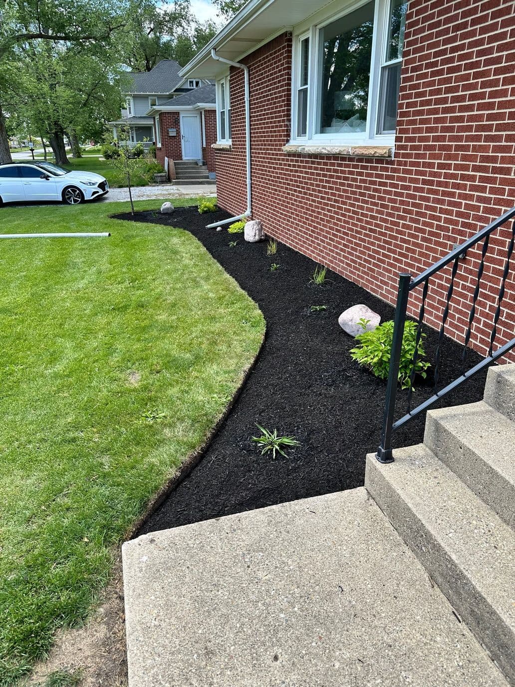 Black mulch landscape featuring plants and rocks alongside a brick house and concrete steps.